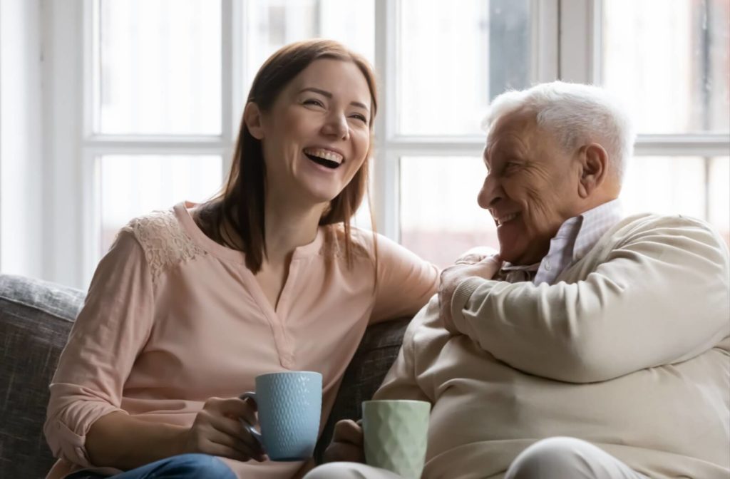 An adult child laughing on the couch while drinking tea with her older father.