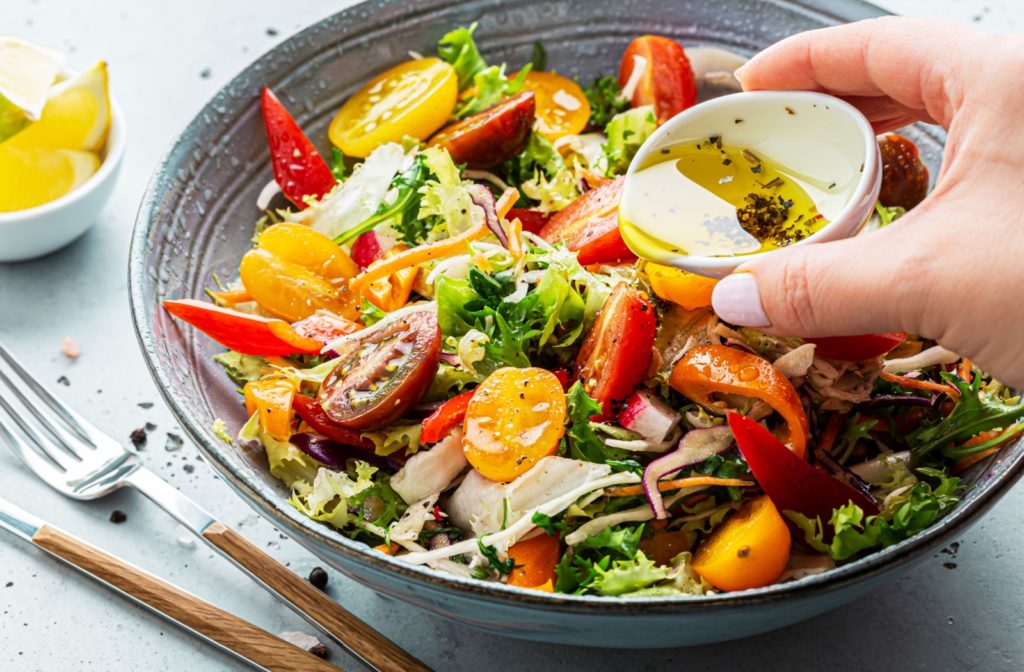 A close-up image of a hand pouring olive oil dressing with herbs over a bowl of salad.