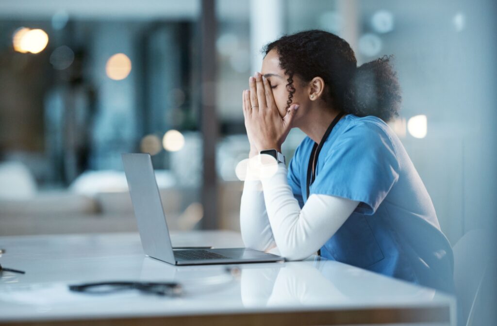 An exhausted young adult in scrubs presses their hands to their face to find relief while trying to do work after caregiving.