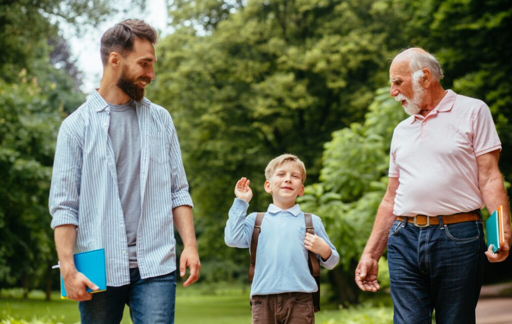 A senior is visited by their adult child and by their young grandchild. They stroll through a green park and enjoy the sunshine.