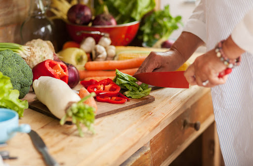 a close up image of a senior's hands chopping lettuce. The cutting board is surrounded by vegetables including daikon, peppers, broccoli and onions