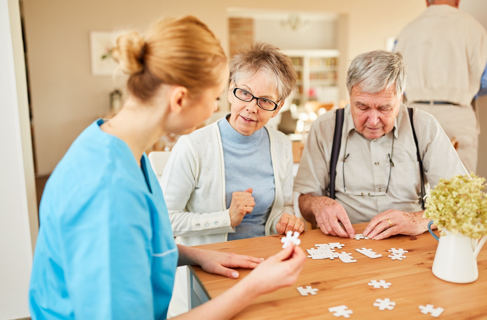 A pair of older adults trying to solve a jigsaw puzzle with the assistance of a memory care staff.