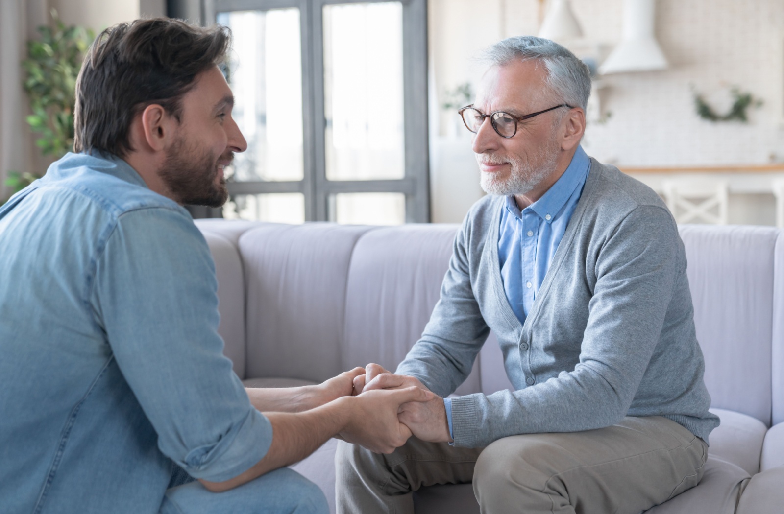 A person holding hands with an older parent while having a heartfelt conversation on a couch.