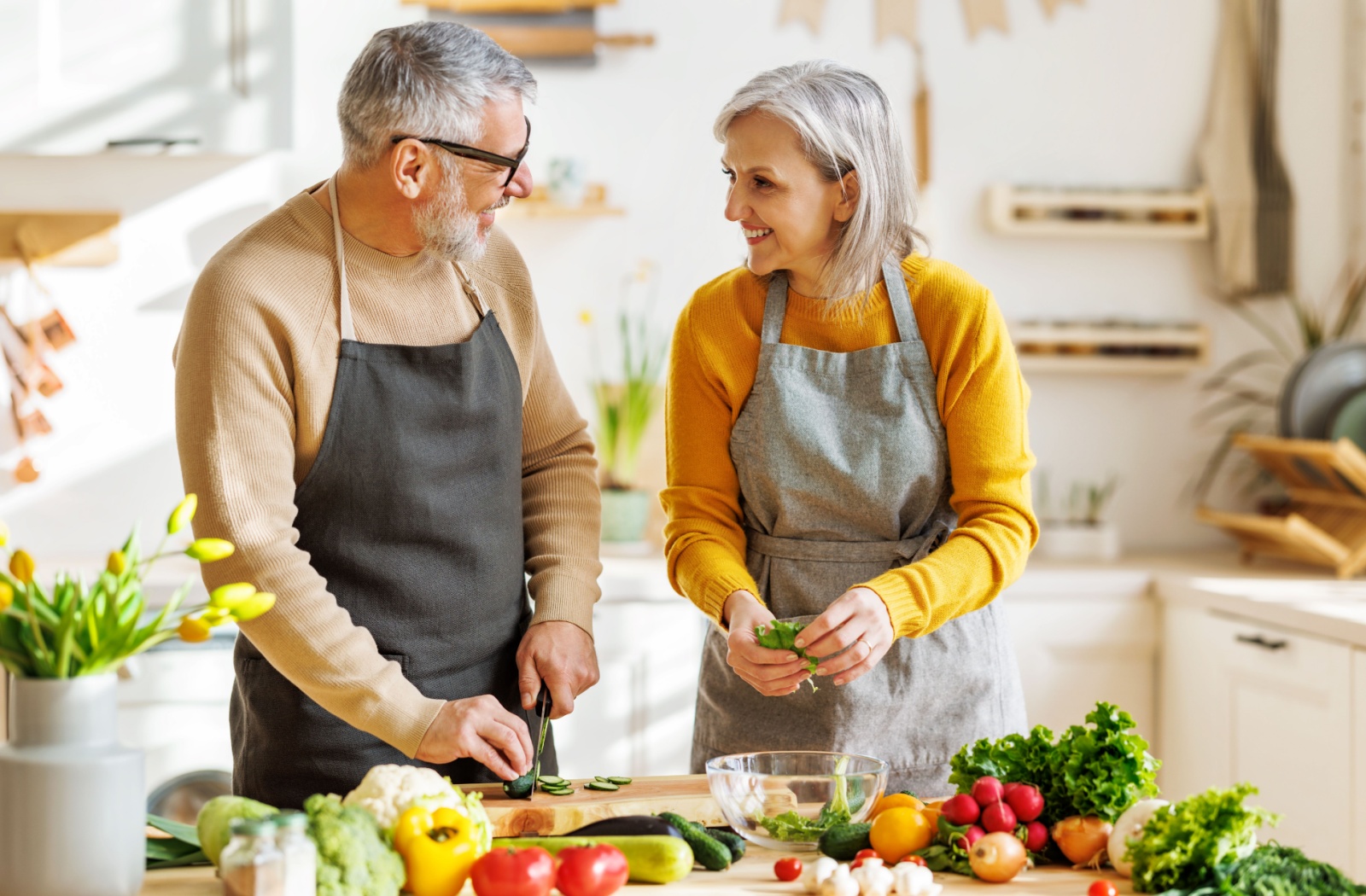 A happy older couple wearing aprons and chopping vegetables in a kitchen.