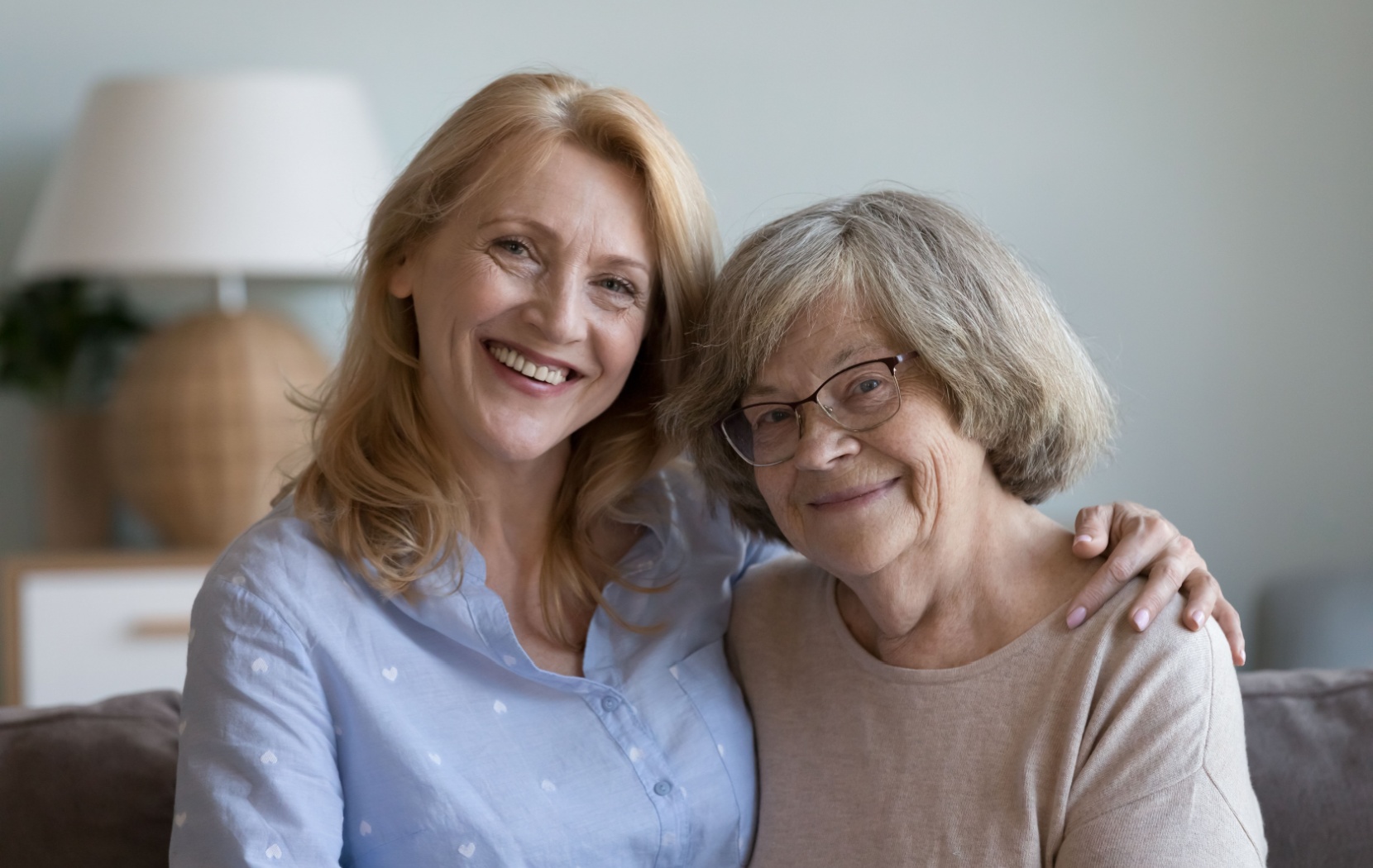 An adult child wraps their arm around their aging parent's shoulders. Both of them smile as they enjoy each other's company.