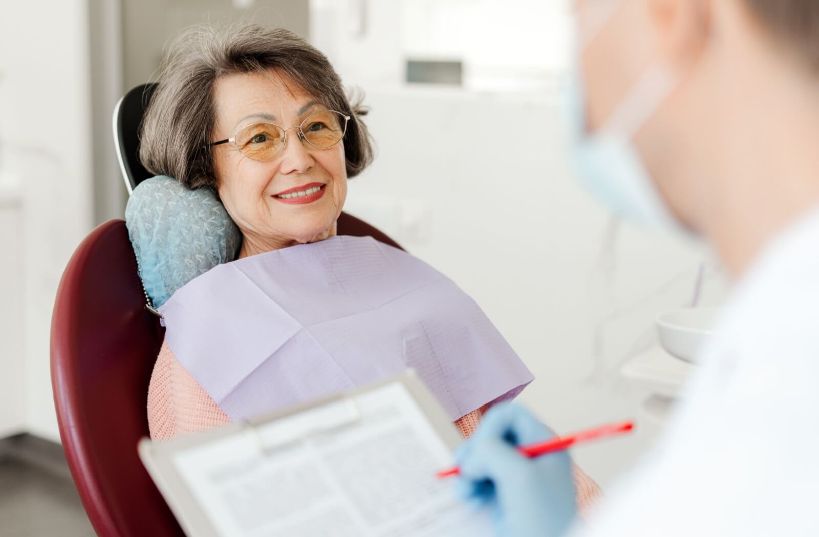 A senior sits back in a dentists’ chair, talking to their dentist about their oral health care needs
