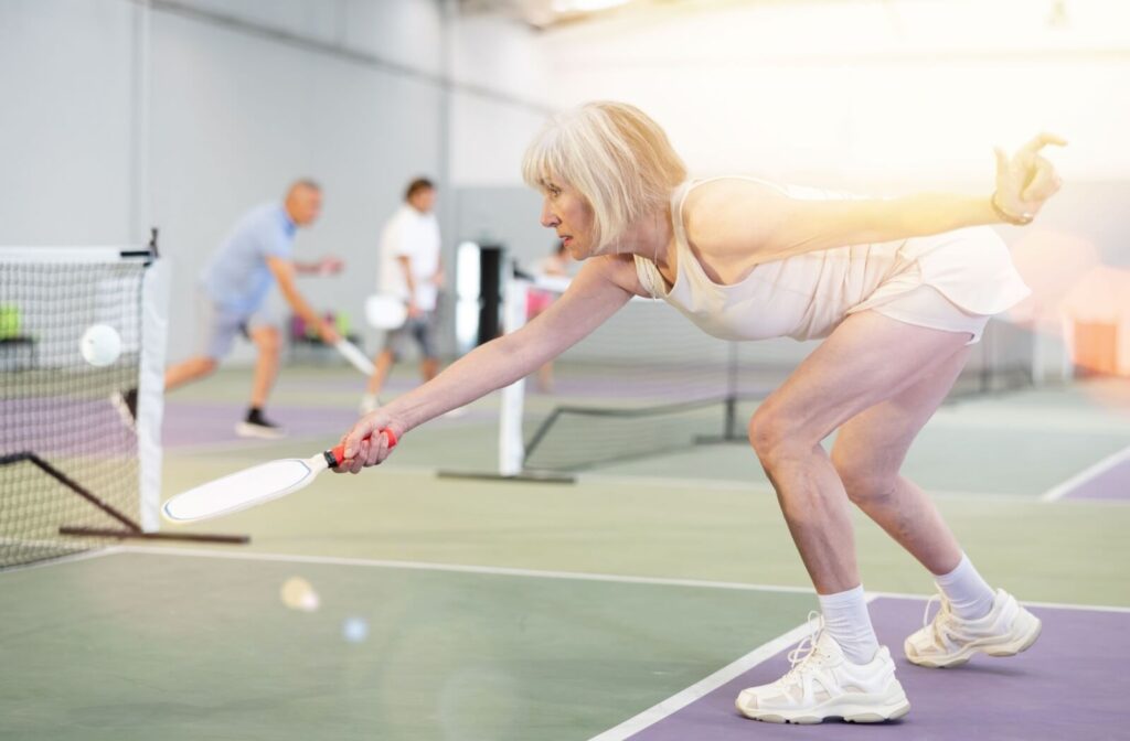 A senior bends forward to hit a wiffle ball while playing pickleball on an indoor court with other seniors