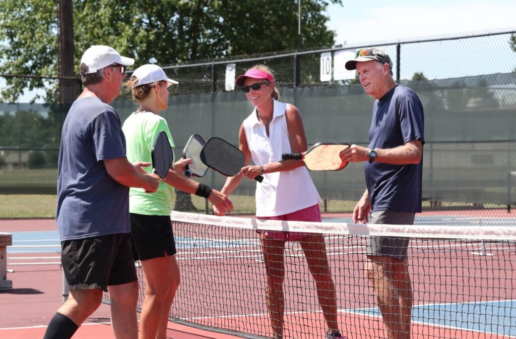Two senior couples talk at the net on an outdoor pickleball court to decide how they’re going to split up teams for a game