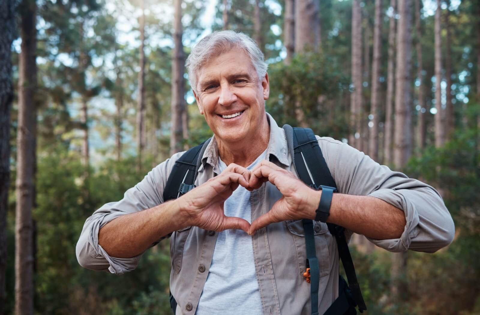 A senior holds their hands in the shape of a heart over their chest while outdoors hiking for exercise to stay healthy