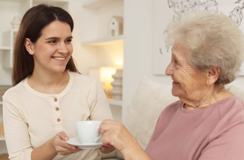 Smiling young woman handing a cup of tea to a senior woman while sitting together in a bright living room