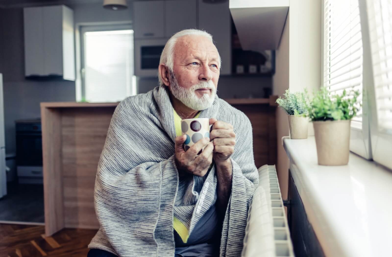Senior wrapped in blanket holding a mug by the window during flu season